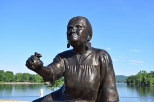 New Sculpture in La Crosse Honors Ho-Chunk Healer Betsy Thunder - Common Good News Bronze statue of a woman kneeling with one arm extended, situated outdoors by a river with trees and a lamppost in the background.