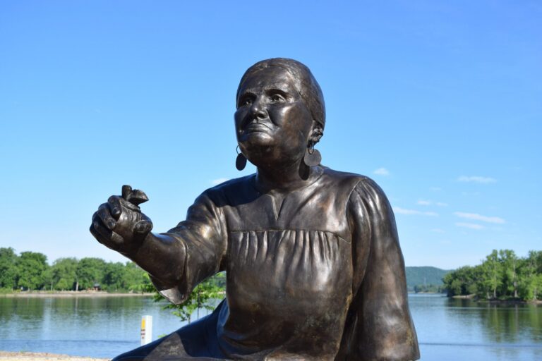 Bronze statue of a woman kneeling with one arm extended, situated outdoors by a river with trees and a lamppost in the background.