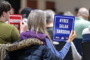 A person seated at an event holds a sign that reads #Free Salah Sarsour alongside other participants holding similar protest signs.