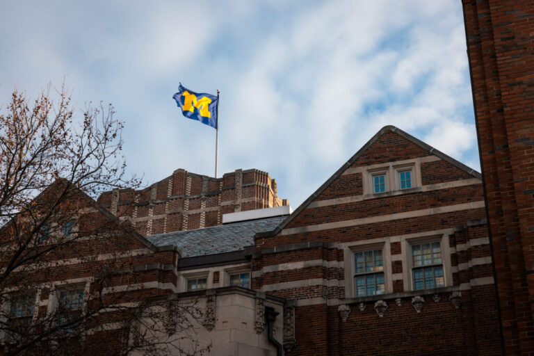 A U-M flag flies atop a building with blue sky and white clouds