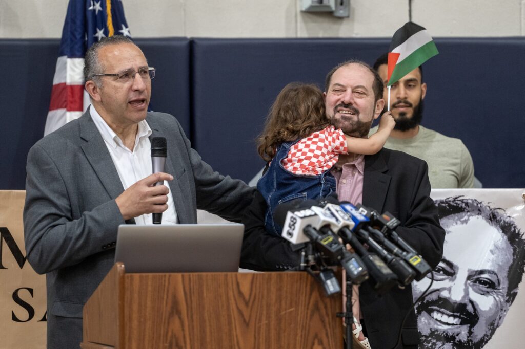 A man speaks at a podium with a microphone, raising one finger, surrounded by adults and children at an indoor event. Microphones and a banner are visible.