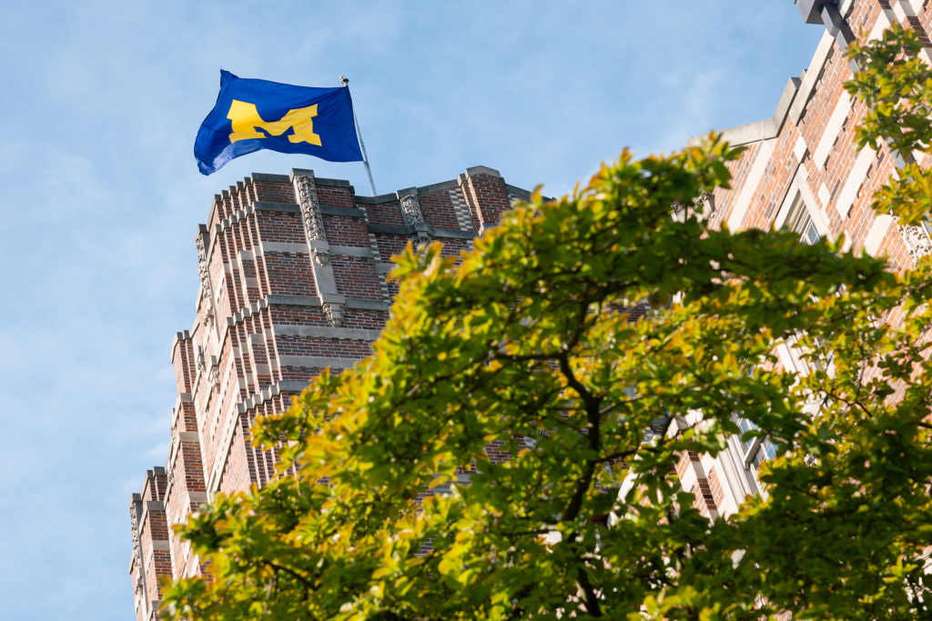 A University of Michigan flag flies atop a brick structure with a tree in the foreground and blue skies