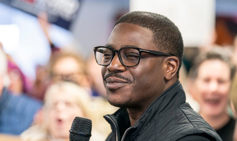 A man with glasses and a black vest holds a microphone and speaks to a crowd. Several blurred faces are visible in the background.
