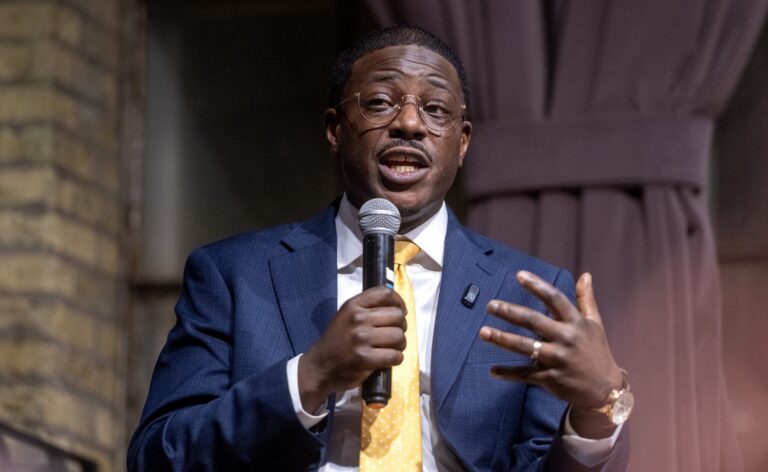 A man in a blue suit and yellow tie speaks into a microphone while gesturing with his hand during an indoor event.