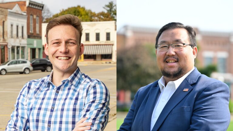 Two men standing outdoors in front of buildings, each smiling with arms crossed. The man on the left wears a plaid shirt; the man on the right wears a blue blazer and glasses.