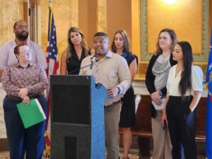 A man speaks at a podium while six people stand behind him in a formal room with an American flag in the background.