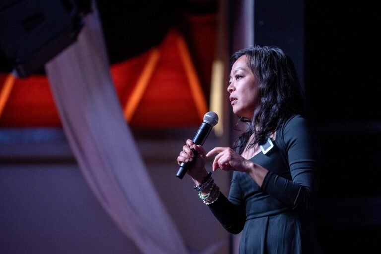 A woman in a dark dress holds a microphone and speaks on stage, with a draped fabric and lights in the background.