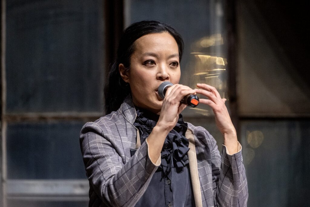 Person in a plaid suit jacket speaks into a microphone, gesturing with one hand, against an industrial-style window background.