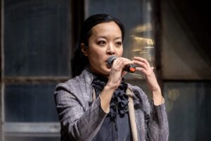 Person in a plaid suit jacket speaks into a microphone, gesturing with one hand, against an industrial-style window background.