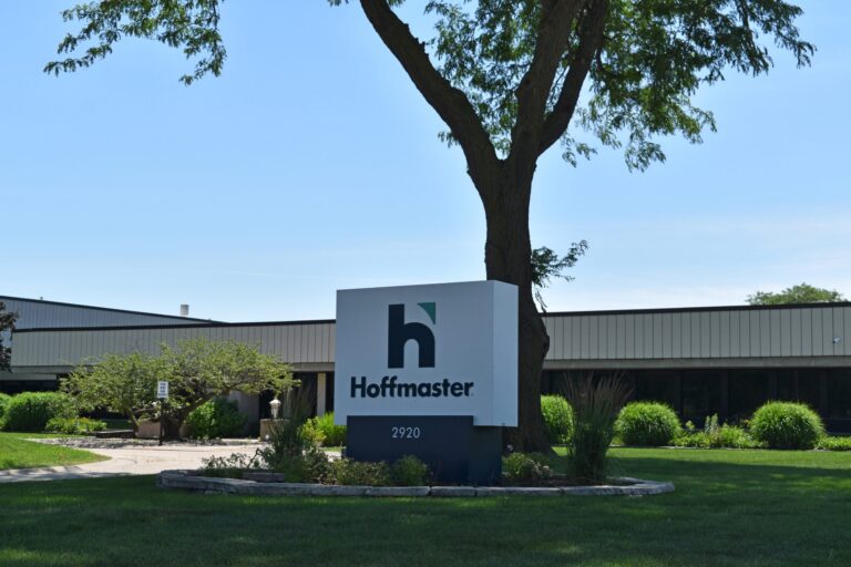 A sign reading Hoffmaster stands in front of a single-story office building with trees and landscaping on a sunny day.