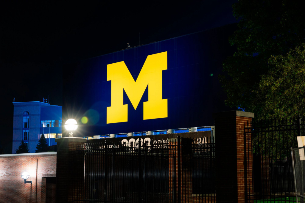 A yellow block M is lit up on a scoreboard at a football stadium at night