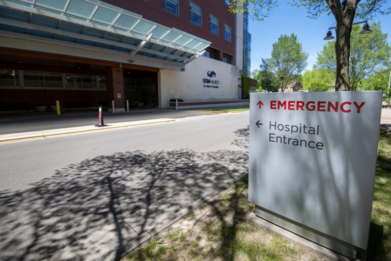 A hospital entrance sign directs to the emergency room and main hospital entrance outside a medical facility on a sunny day.