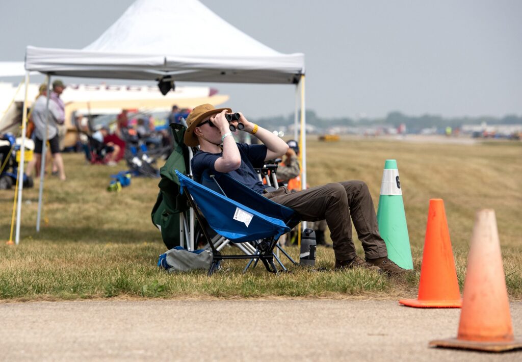 Several attendees sit in lawn chairs below the wings of a large silver airplane.