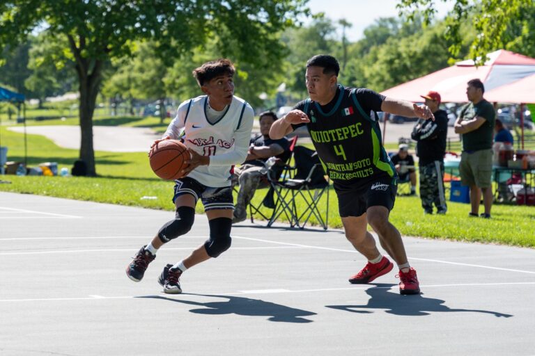 People watch a basketball game outdoors from under a tent; players in purple and black compete on the court, with trees and a blue sky in the background.