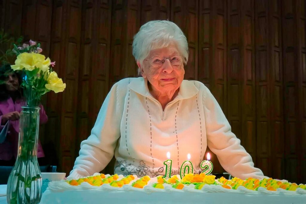 An elderly woman stands behind a cake with lit candles showing 102 and yellow decorations, with a vase of flowers nearby.