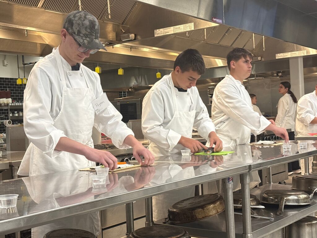 Students and an instructor assemble model vehicles at a workbench, using instructions and wearing safety goggles and gloves.