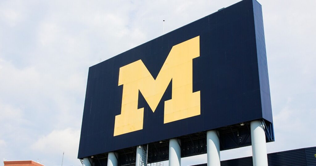 Large blue sign with a yellow Michigan 'M' logo against a cloudy sky.