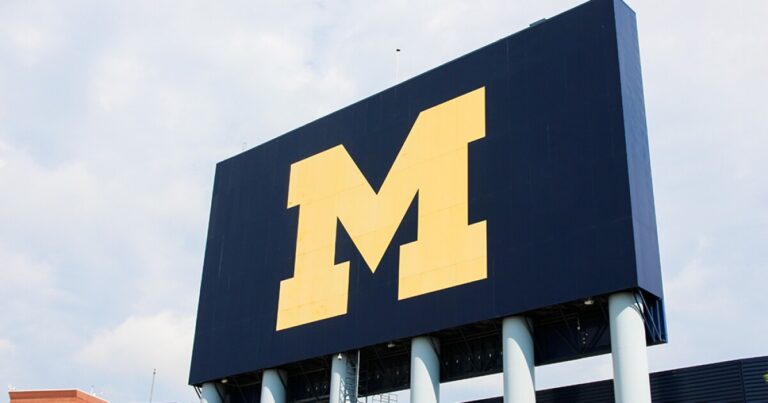 Large blue sign with a yellow Michigan 'M' logo against a cloudy sky.