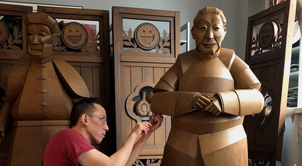 A woman looks at a wooden sculpture of a seated figure displayed on a white pedestal in an art gallery.