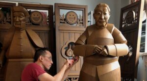 Wisconsin Artist Warren King Crafts Life-Sized Sculptures from Cardboardembedhttpswwwyoutubecomwatchv=J9oSoj8oI-Yembedembedhttpswwwyoutubecomwatchv=iqpnGTKwES0embed - Common Good News A woman looks at a wooden sculpture of a seated figure displayed on a white pedestal in an art gallery.