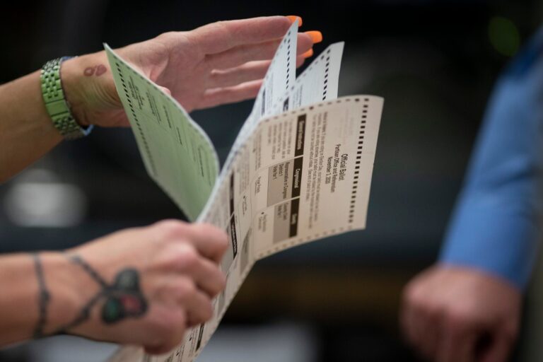 A person holds and examines a folded paper ballot, with visible tattoos and an orange manicure, during an election process.
