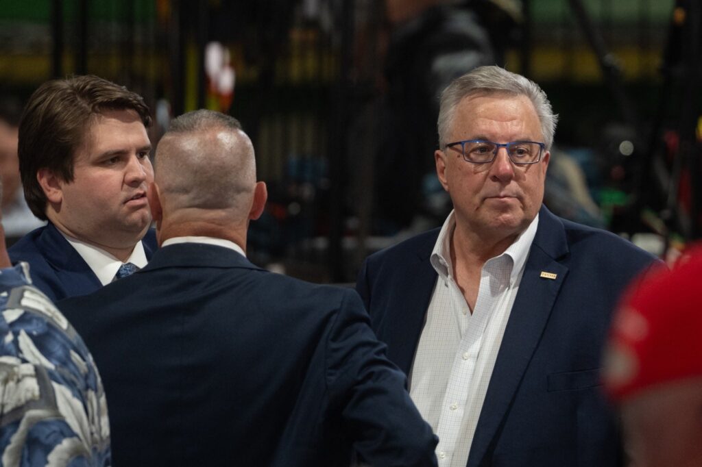 Three men in suits stand and talk indoors; one faces the camera while the others are turned away or in profile. The background is blurred and busy.