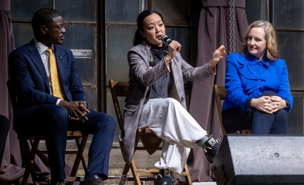 Three people sit on stage chairs; the person in the center speaks into a microphone while the others listen attentively.