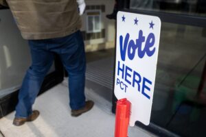 A person enters a building next to a sign that reads Vote Here PCT with blue text and stars.