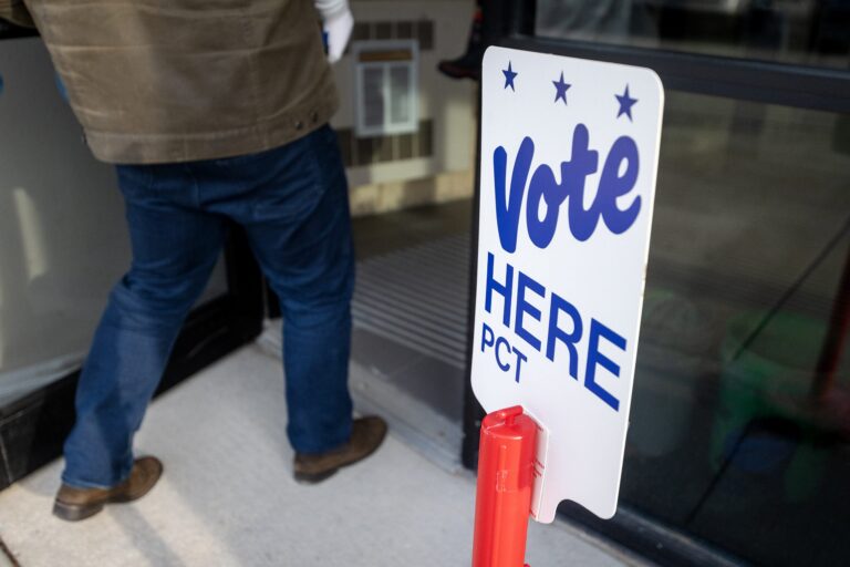 A person enters a building next to a sign that reads Vote Here PCT with blue text and stars.