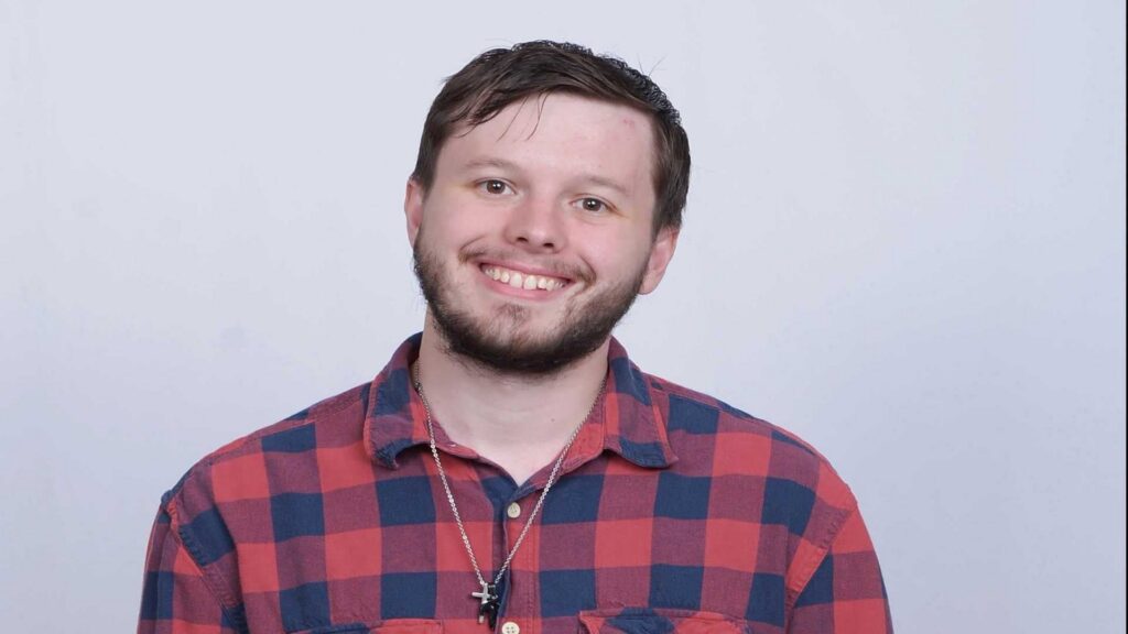 A person with short brown hair and a beard is smiling, wearing a red and blue plaid shirt and a necklace with a cross, against a plain light background.