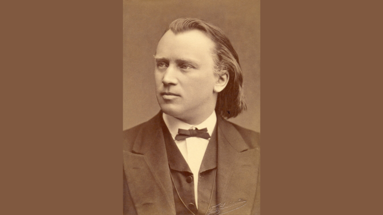 Brahms Symphony Journey From Beethovens Shadow to MSOs Stage - Common Good News An older man in a black shirt stands indoors, leaning on a railing and holding a conductors baton, looking to the side with a thoughtful expression.