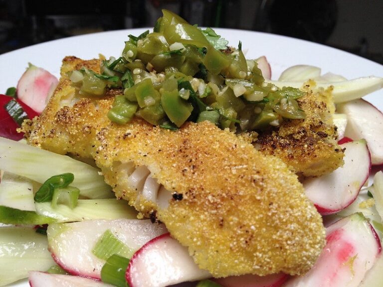 Person preparing whole fish with lemon slices on a cutting board near a sink, olive oil, and a green checkered towel.