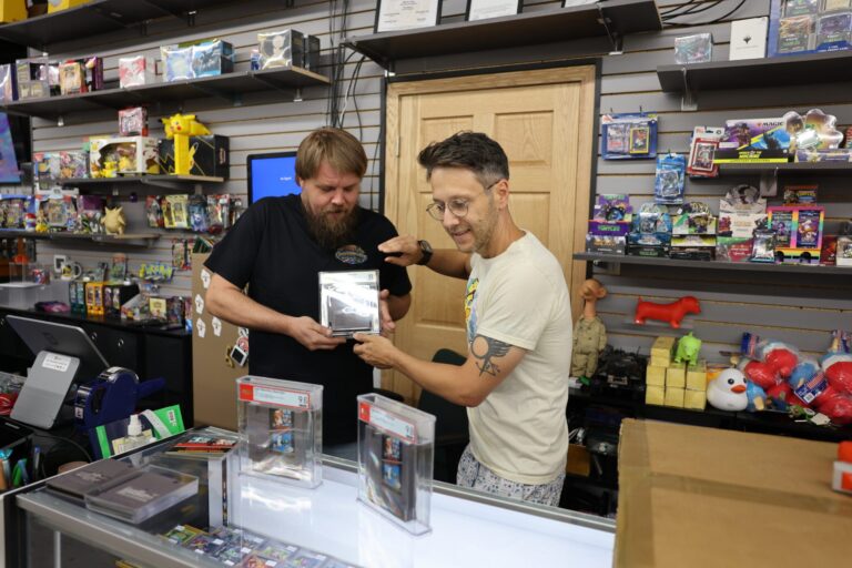 Two men stand behind a store counter filled with collectibles; one man lifts a large cardboard box while the other looks on and smiles.