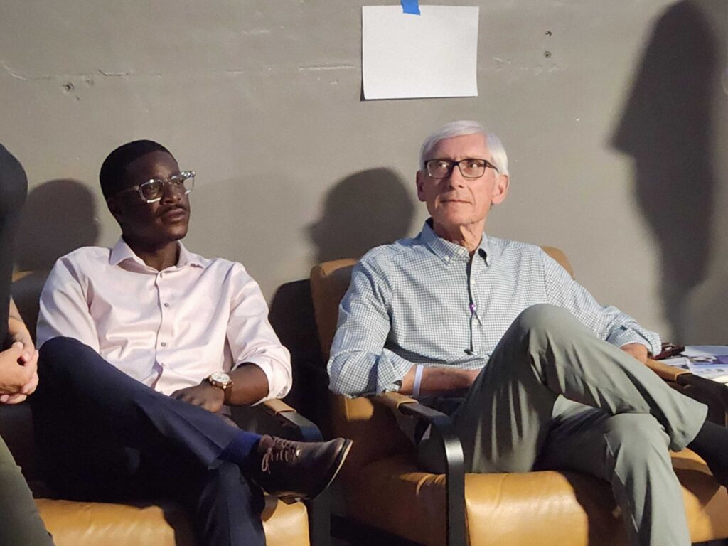 Two men sit side by side in brown chairs against a gray wall, looking forward. One wears a white shirt and glasses, the other wears a light blue shirt and glasses.