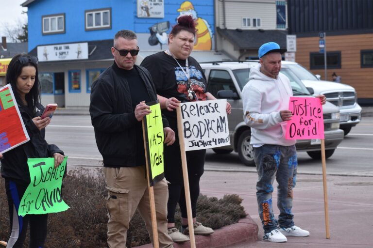 Four people stand on a sidewalk holding protest signs, including messages like Release Bodycam and Justice for David, with cars and buildings in the background.