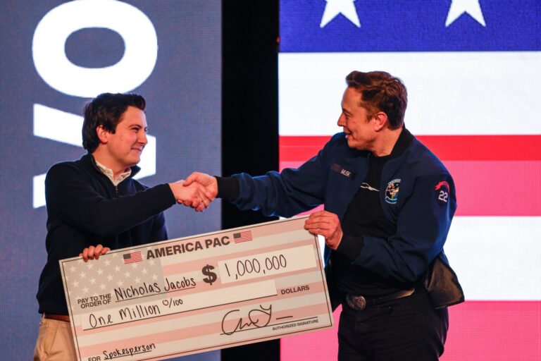 Two men shaking hands on stage, one holding a large novelty check for one million dollars with Nicholas Jacobs written on it, against a backdrop of the American flag.