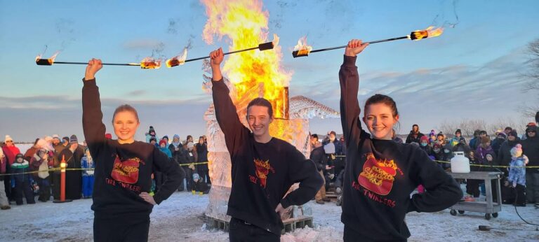 Three performers hold flaming batons in front of a large outdoor fire, with a crowd watching in the background on a snowy day.