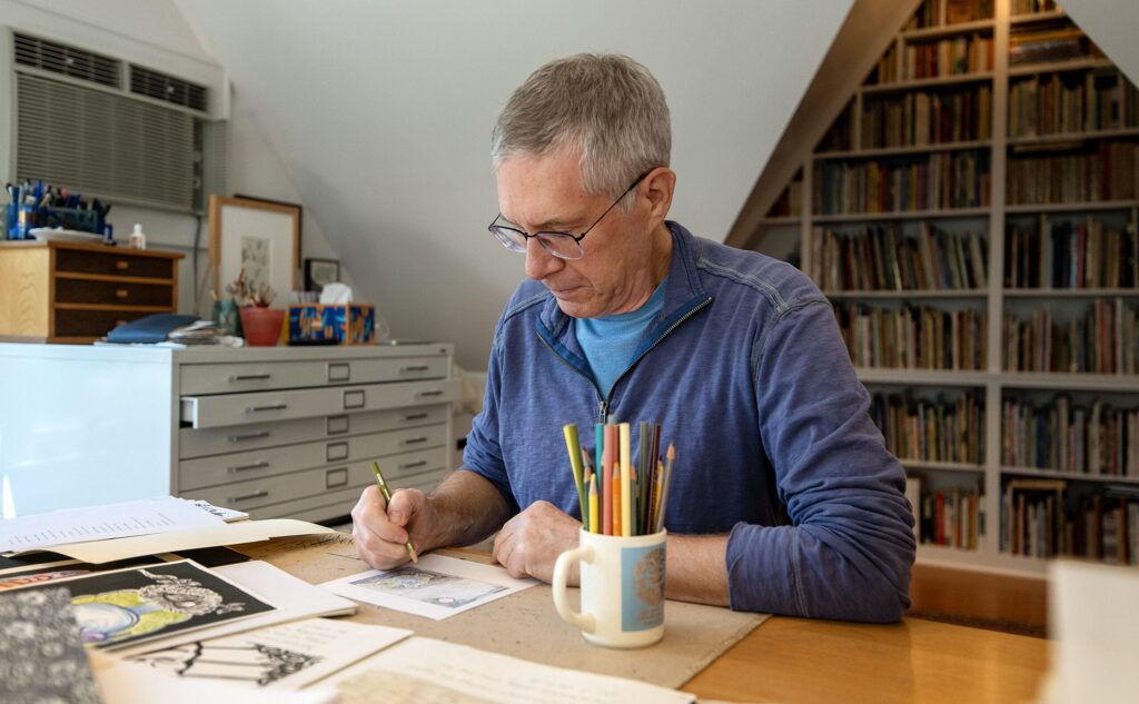 A man with gray hair and glasses draws at a desk with papers, art supplies, and a mug of colored pencils, in a room with shelves full of books.