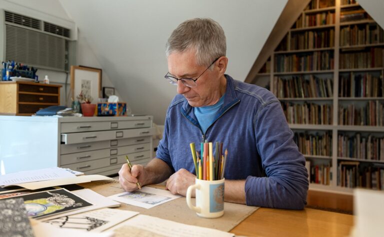 A man with gray hair and glasses draws at a desk with papers, art supplies, and a mug of colored pencils, in a room with shelves full of books.