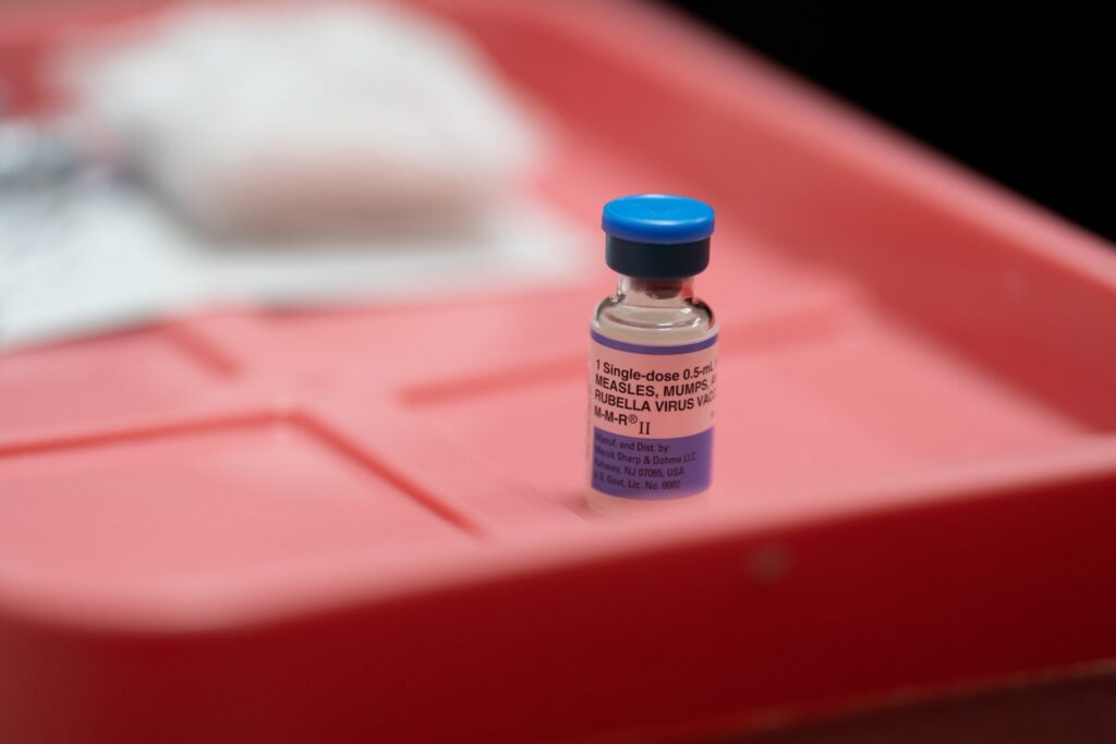 A small vial with a blue cap labeled for measles, mumps, and rubella vaccine sits on a red plastic surface.