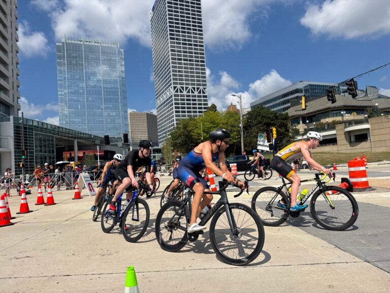 Cyclists wearing helmets race on a city street surrounded by tall buildings, traffic cones, and barriers under a partly cloudy sky.