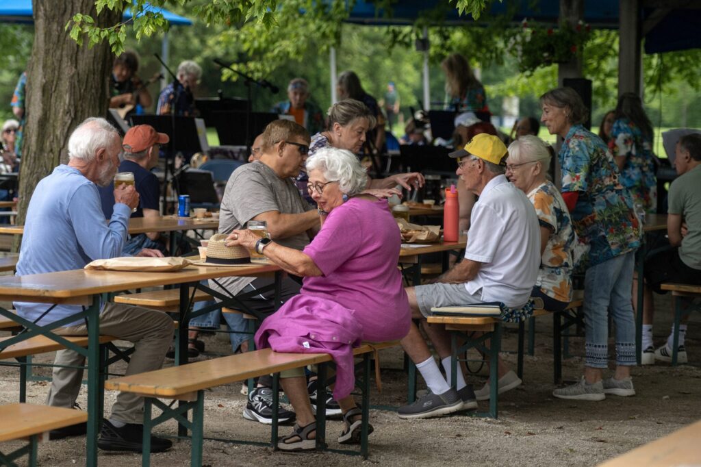 People gather at an outdoor Biergarten with blue umbrellas and tables, surrounded by greenery. A man walks toward the seating area, and bicycles are parked nearby.