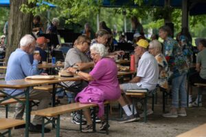 Milwaukees Beer Gardens A Decade-Long Summer Social Phenomenon - Common Good News People gather at an outdoor Biergarten with blue umbrellas and tables, surrounded by greenery. A man walks toward the seating area, and bicycles are parked nearby.