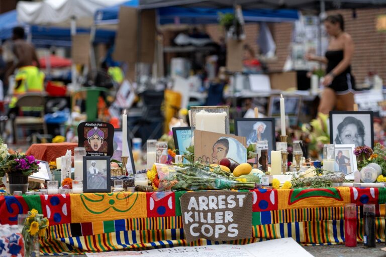 A memorial table with photos, candles, flowers, and a sign reading “ARREST KILLER COPS” at a public gathering with people and tents in the background.