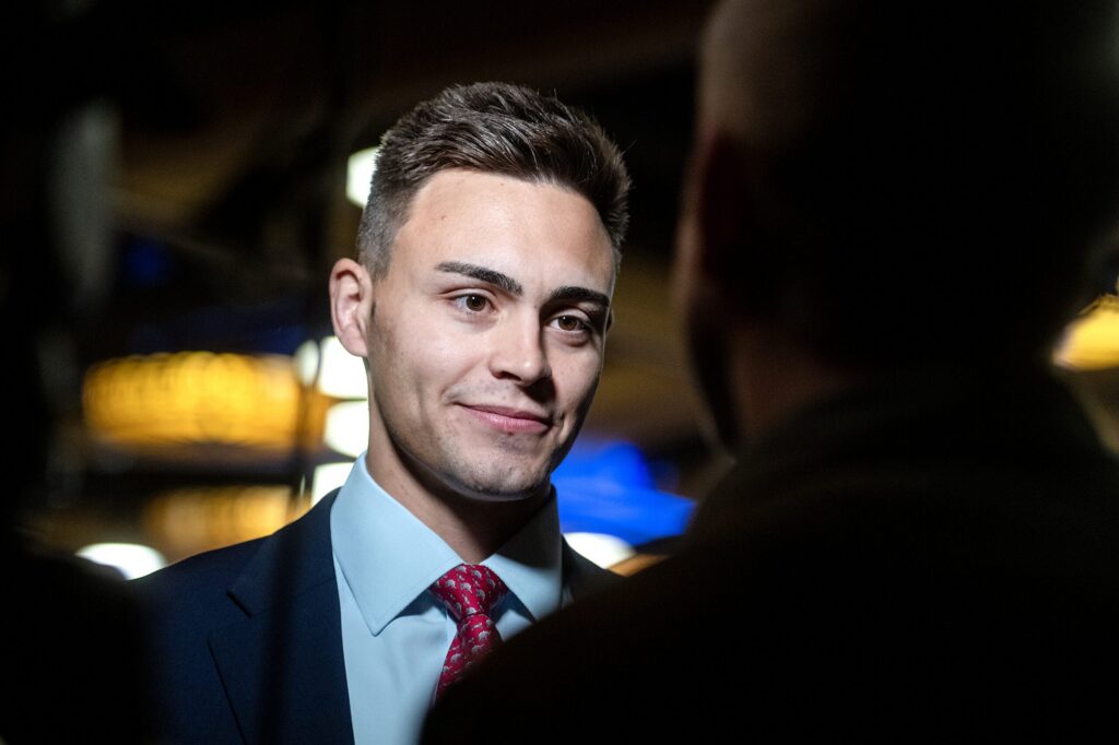 A young man in a suit and tie speaks to another person in a dimly lit setting with blurred lights in the background.