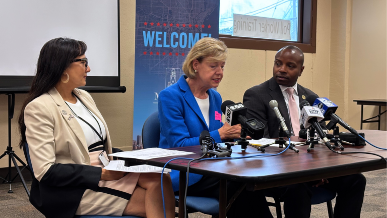 Three people sit at a table with microphones during a press conference. A Welcome sign is in the background. One woman speaks while the others listen attentively.