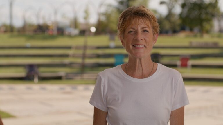 A woman with short hair in a white t-shirt stands outdoors, smiling at the camera, with a grassy park and benches in the background.