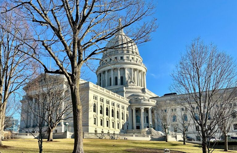 A large, domed government building with columns and a prominent staircase, surrounded by leafless trees and a grassy lawn under a clear blue sky.