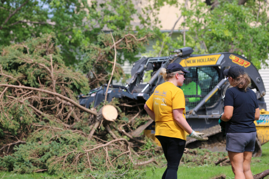 Sisters watch as a skid-steer loader pulls a tree in Highmore, South Dakota, on June 29, 2026, after a severe storm that morning. The Federal Emergency Management Agency announced it will reimburse eight South Dakota counties and three tribes for storm-related infrastructure fixes. (Photo by Meghan O’Brien/South Dakota Searchlight)