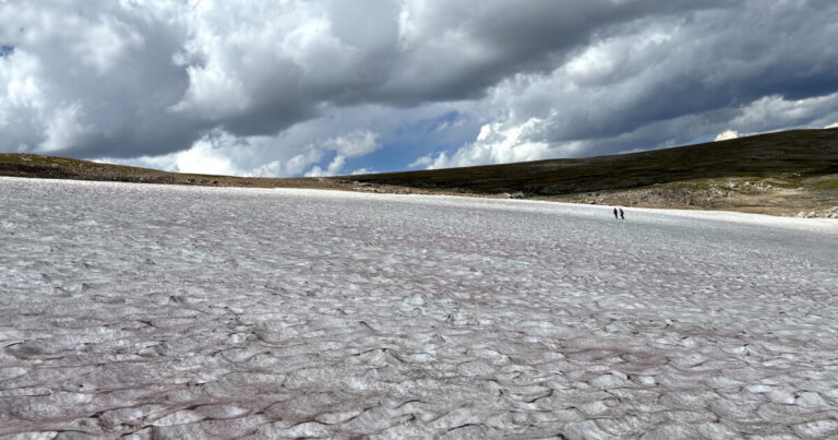 Two researchers use a power drill in rocky ground beside a high-elevation ice patch.